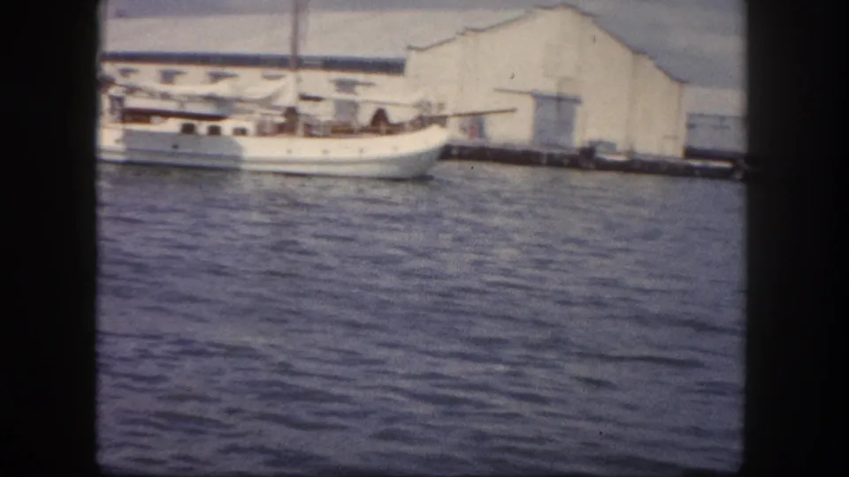 8mm frame circa 1960, Tampa harbor: a white sailing vessel tied up alongside a large white warehouse shed, viewed from water level with dark porthole vignetting.