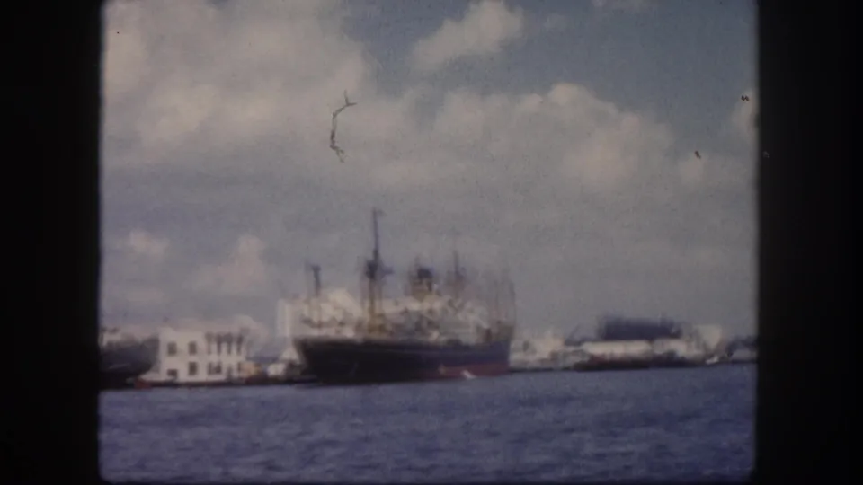 8mm frame circa 1960, Tampa harbor: large cargo freighter with loading cranes moored at a pier, low dock buildings behind, cumulus clouds above; film scratch visible in upper frame.