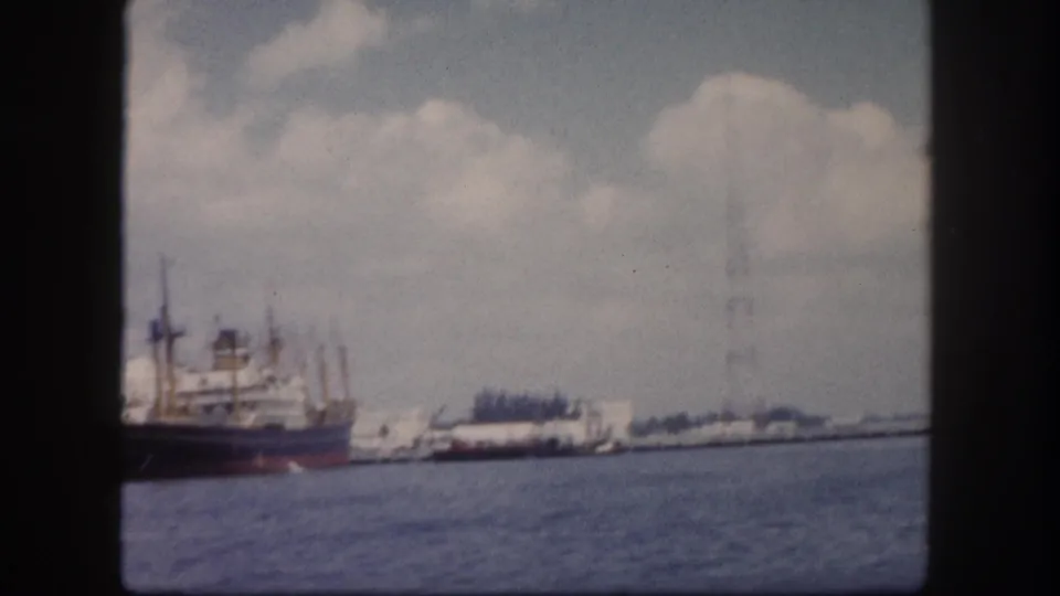 8mm frame circa 1960, Tampa harbor: same large cargo freighter with dark red-brown hull and loading cranes, waterfront buildings visible behind under cumulus clouds, angle shifted as boat passes.