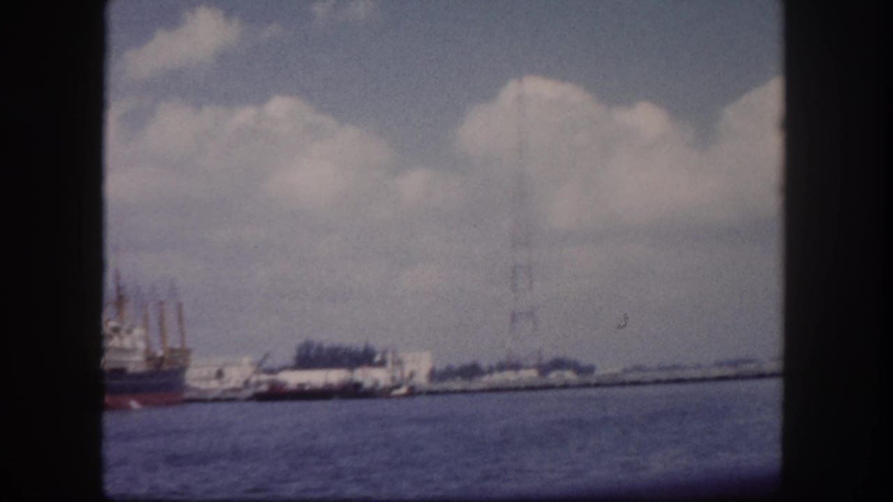 Harbor view from a boat showing a docked cargo ship with a red hull and a tall steel lattice tower, with choppy water in the foreground.
