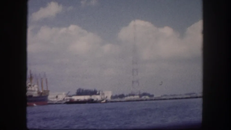 8mm home movie frame circa 1960 showing Tampa harbor: a red-hulled cargo ship at dock, a tall steel lattice tower, and choppy foreground water under cumulus clouds.
