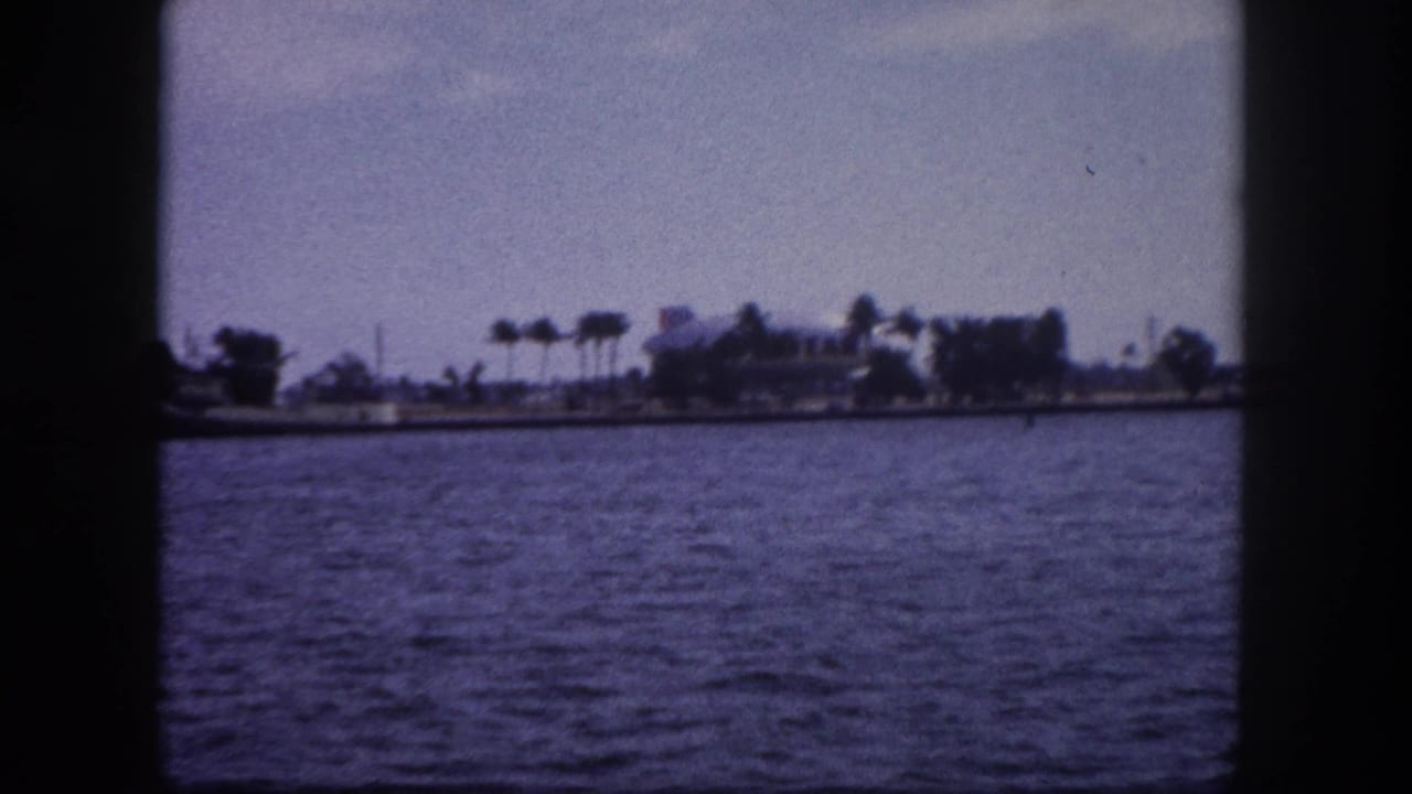 8mm home movie frame circa 1960 showing open Tampa Bay water with a palm-lined shoreline and scattered low buildings in the middle distance.