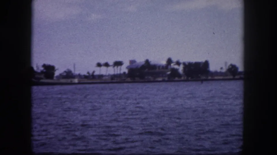 8mm home movie frame circa 1960 showing open Tampa Bay water with a palm-lined shoreline and scattered low buildings in the middle distance.