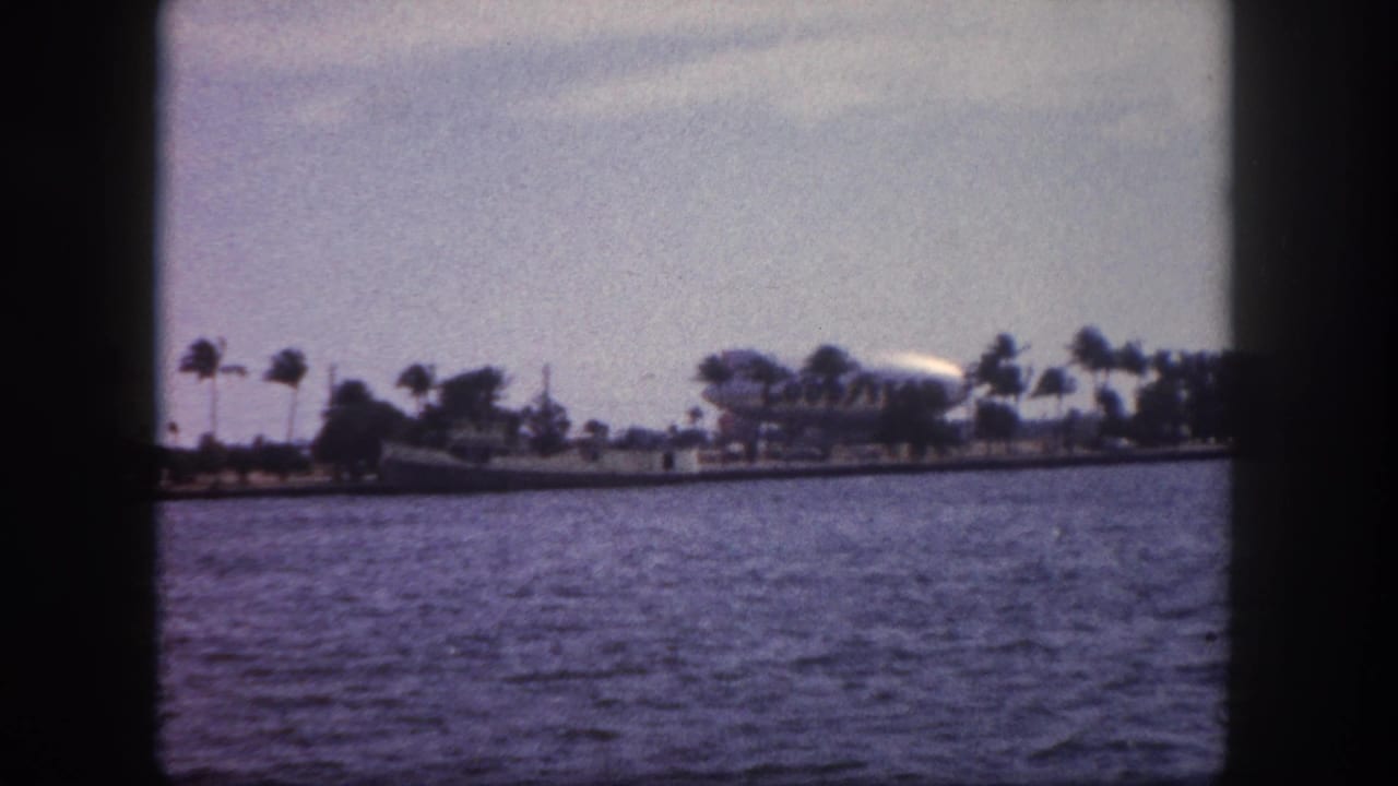 8mm film frame showing Tampa Bay waterfront with palm trees and a small white airship or blimp visible on the distant shore.