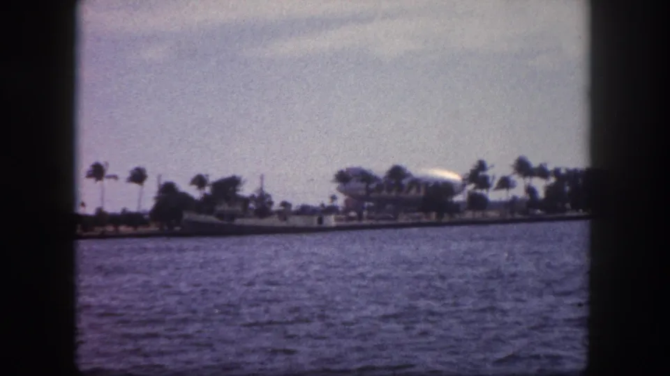 8mm home movie, 1960 Tampa Florida: open waterway with palm-lined shoreline and a large white airship or blimp visible in the right background.