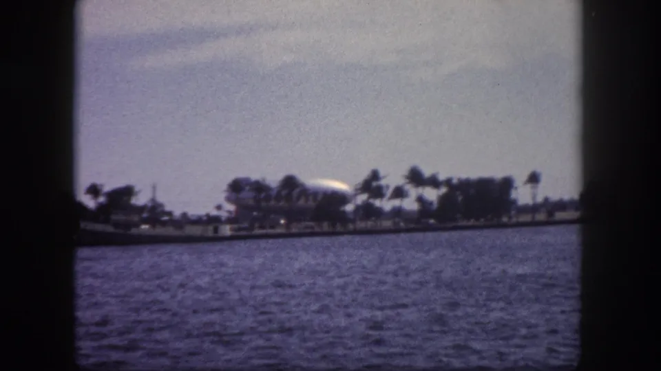 8mm home movie, 1960 Tampa Florida: large white airship grounded near the waterfront, palm trees and low buildings visible across the bay from a moving boat.