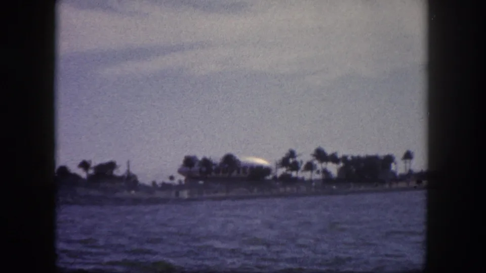 8mm home movie, 1960 Tampa Florida: boat-level pan of waterfront with palm trees, low structures, and a large white airship at the right edge of the distant shore.