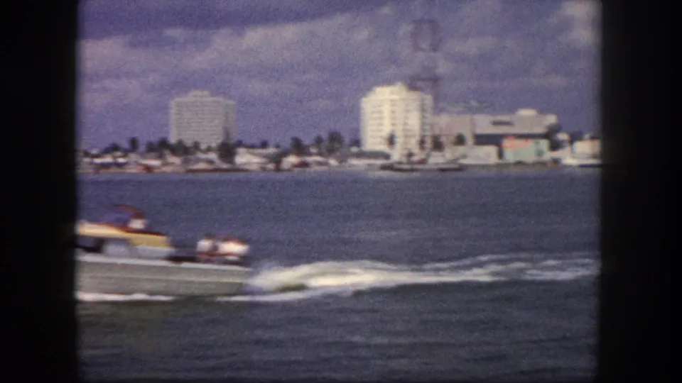 Motorboat crossing Tampa waterfront circa 1960; mid-rise commercial buildings and palm trees along the distant shoreline, choppy water in the foreground.