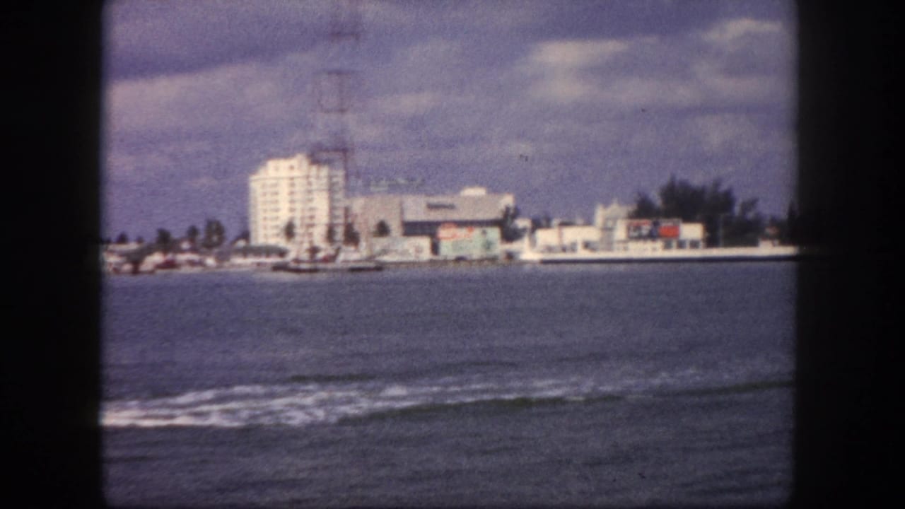 8mm pan frame from water showing Tampa waterfront with white mid-rise building, tall lattice tower, palm trees, low commercial buildings, and choppy foreground water.