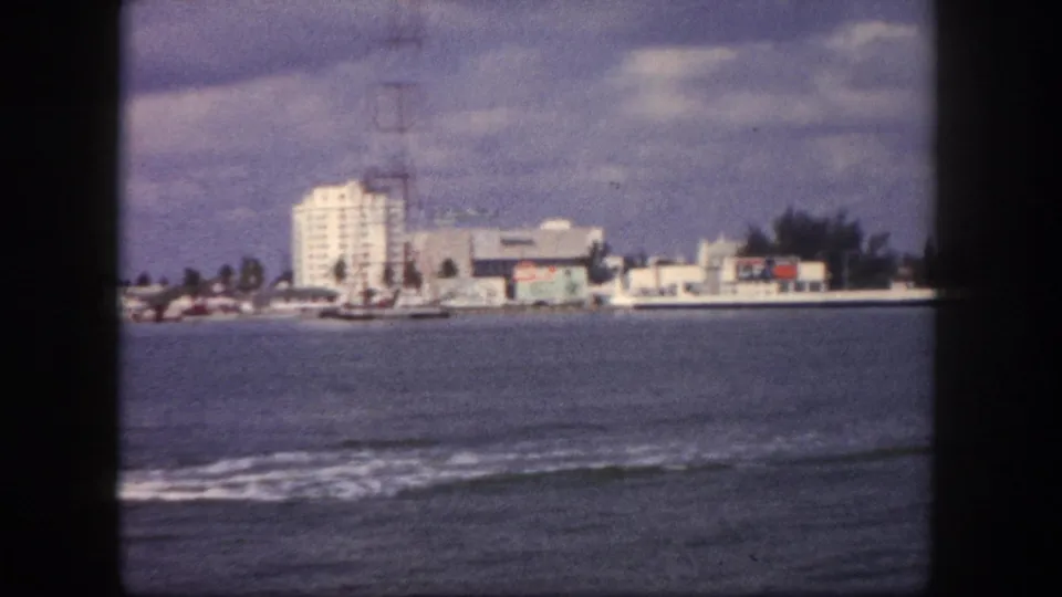 8mm home movie frame, circa 1960, Tampa, Florida waterfront seen from a boat: white multi-story building, tall lattice transmission tower, palm trees, low commercial buildings along shore.