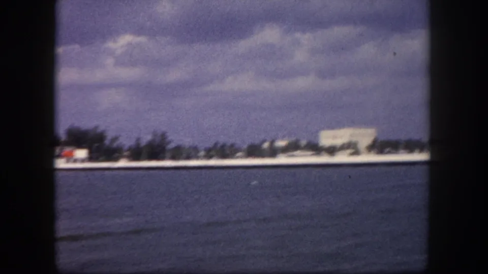 8mm home movie pan frame, circa 1960, Tampa area, Florida: flat waterfront strip with palm trees, low-rise buildings, and open bay water filling the foreground.