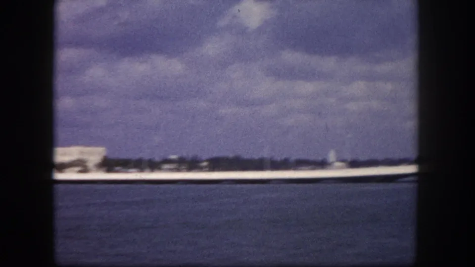 8mm home movie pan frame, circa 1960, Tampa Bay area: broad water expanse dominates, distant flat shoreline at horizon, blue-purple color cast characteristic of aged 8mm film stock.