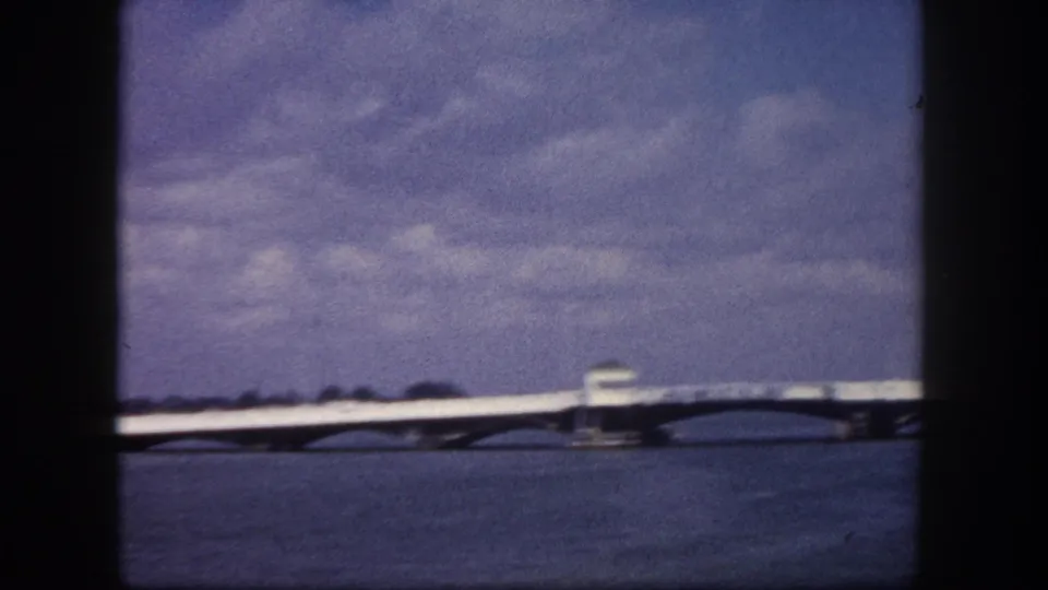 8mm home movie pan frame, circa 1960, Tampa area, Florida: long low-profile bridge or causeway spanning open bay water, distant white building at right, overcast sky with blue-purple aged film tone.
