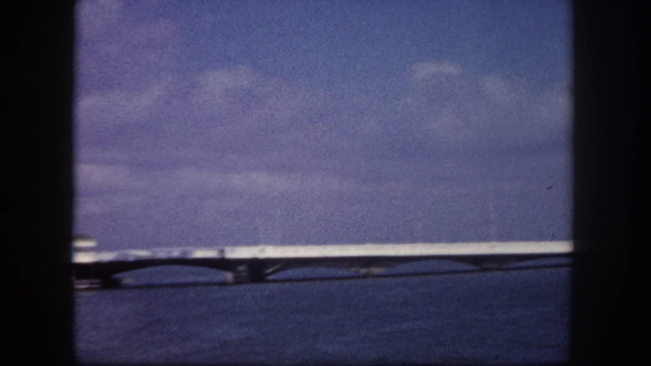 Long causeway bridge spanning calm bay water, blue-purple sky with scattered clouds, viewed from a boat.