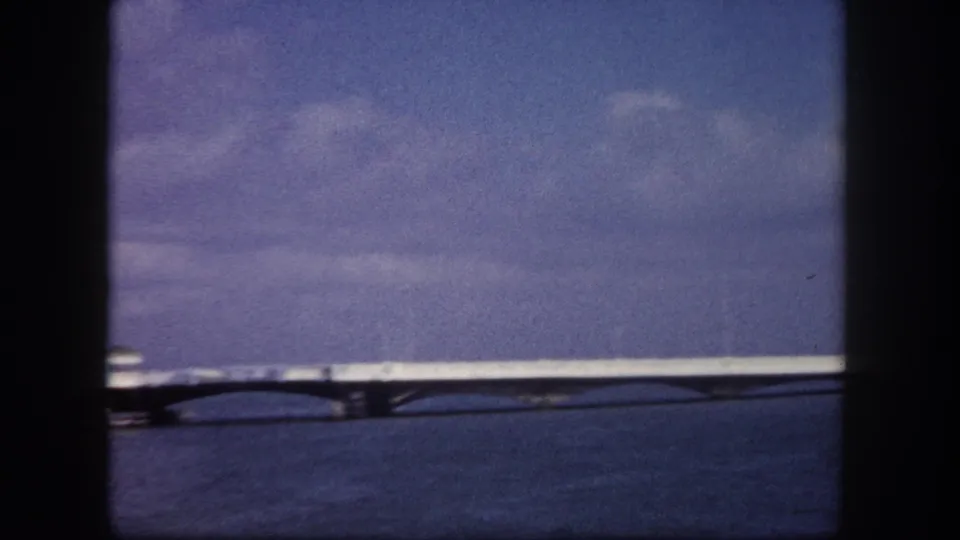 8mm footage circa 1960, Tampa Florida — a long flat bridge crossing calm bay water seen from boat level, blue sky with scattered clouds above.