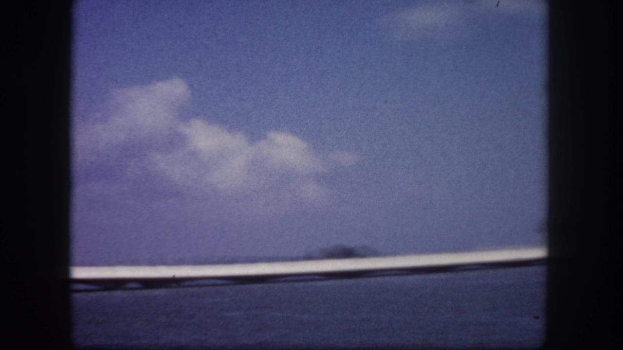 8mm frame circa 1960, Tampa Bay area — causeway bridge extending across calm water with a distant landmass on the right horizon, color-faded film.