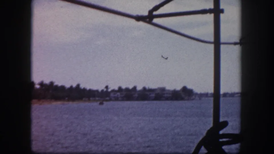 8mm home-movie frame circa 1960, Tampa Florida — warm color-shifted bay water, palm-lined shore, low-rise buildings, and boat framework in upper-right corner.