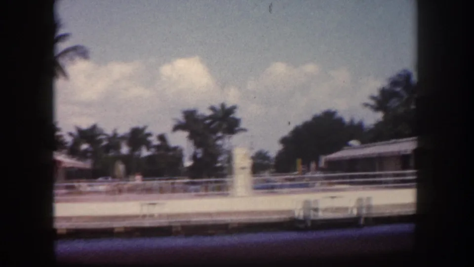 8mm home movie frame from Tampa, Florida, circa 1960: low waterfront building with white fence and palm trees under cumulus clouds, framed through a boat railing.