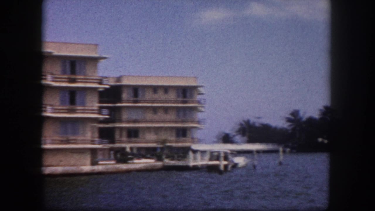 Multi-story tan building at the waterfront with a dock and moored boats; palm trees at the edges against a blue sky.