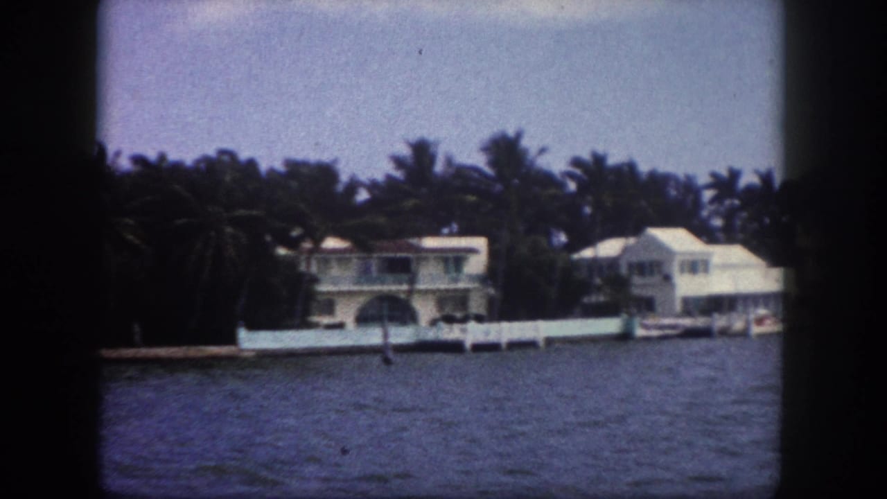 Closer 8mm view of a prominent two-story white estate with arched architectural detail, covered boat slip, and dock fence, Tampa, Florida, circa 1960.