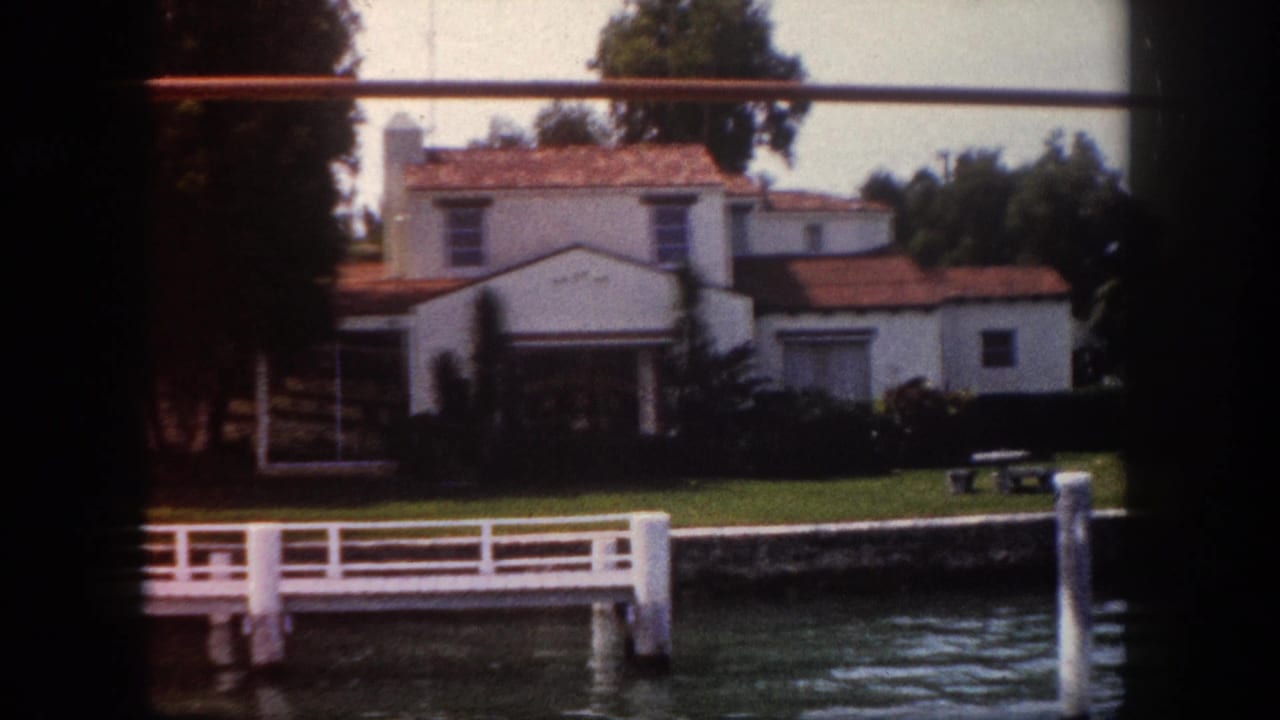 White stucco house with red terracotta tile roof on a waterfront lot, white dock extending into the water, viewed from a boat.