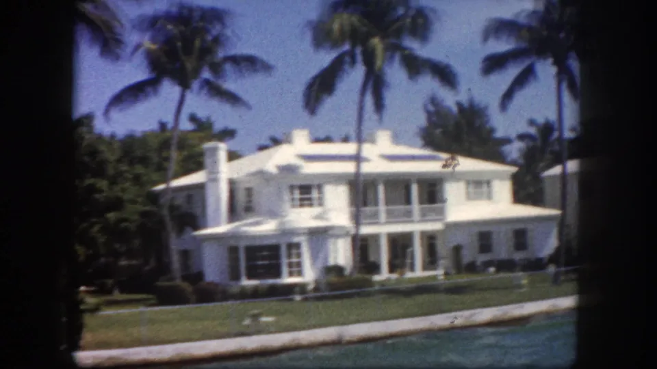 Grand two-story white waterfront mansion with columned upper veranda and manicured lawn, Tampa, Florida area, circa 1960, shot from the water on 8mm home movie film.