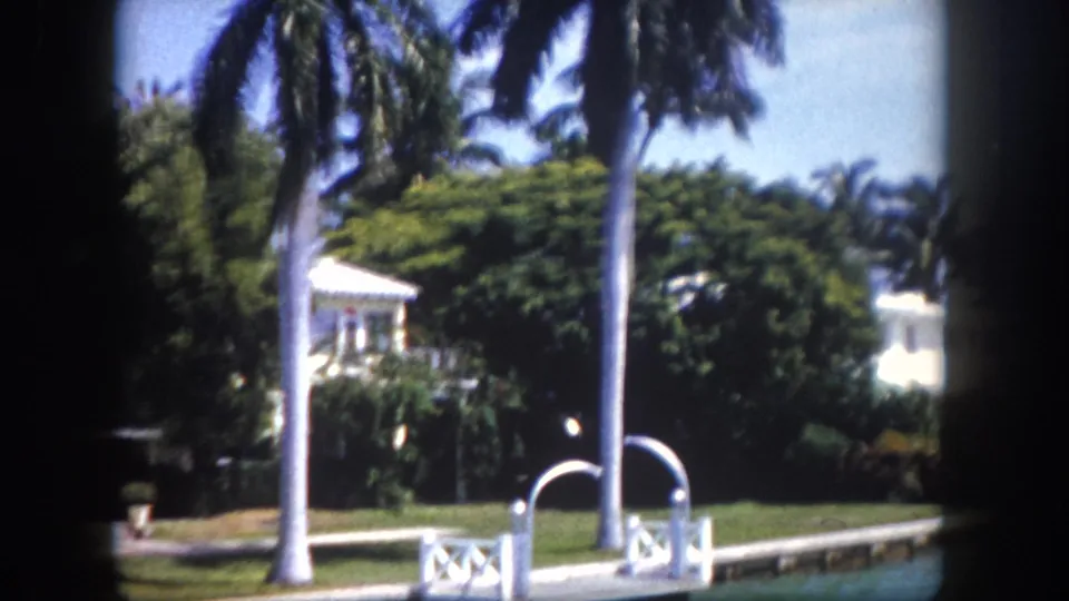 8mm view of a private Florida waterfront dock with white metal rail structures, large palm trunks, and white residential property visible through tropical greenery, circa 1960.