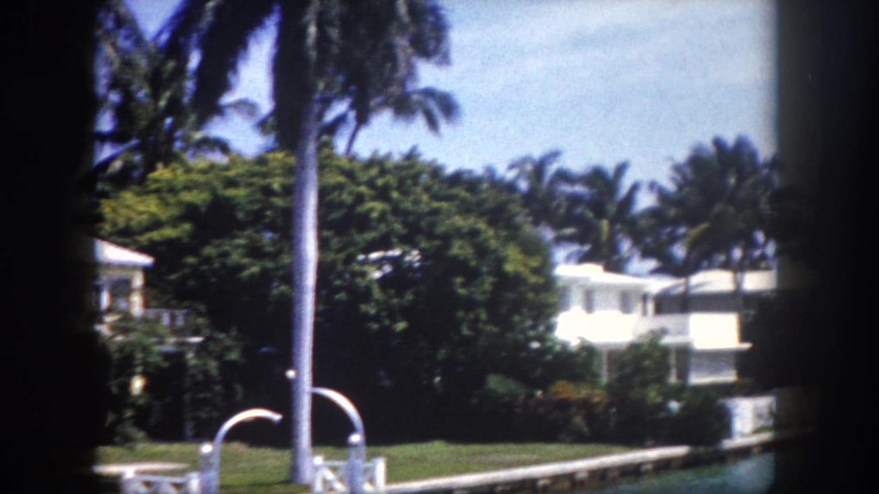 8mm film frame showing white stucco houses and palm trees along a Florida canal, shot from water level with dark vignette edges.