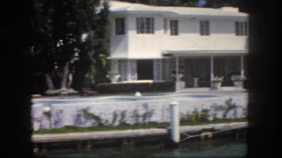 8mm color footage circa 1960, Tampa, Florida: large two-story white stucco house with wraparound covered porch and planters; white dock pilings and calm canal water in foreground.