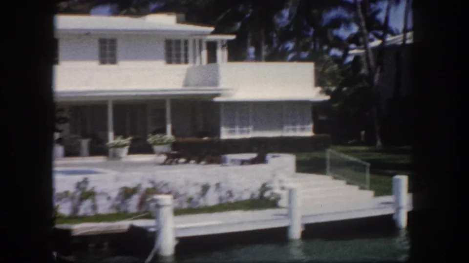 8mm color footage circa 1960, Tampa, Florida: white two-story stucco house with lower colonnaded porch, tropical plantings at waterline, and white dock pilings in foreground.