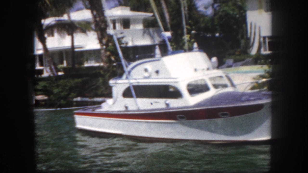 White cabin cruiser with red hull trim on a green waterway, modernist houses and palm trees behind.