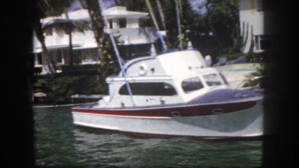 White and red motorboat on a Florida canal waterway with mid-century flat-roofed houses and palm trees in the background, circa 1960 8mm film.