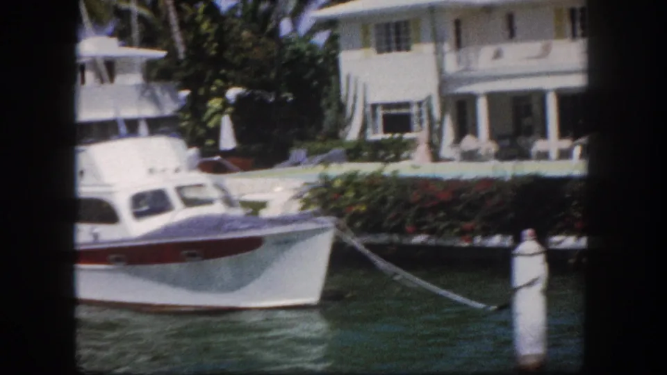 Cabin cruiser moored on a Florida canal with a white columned house, red flowering plants, and palm trees visible, circa 1960 8mm film.