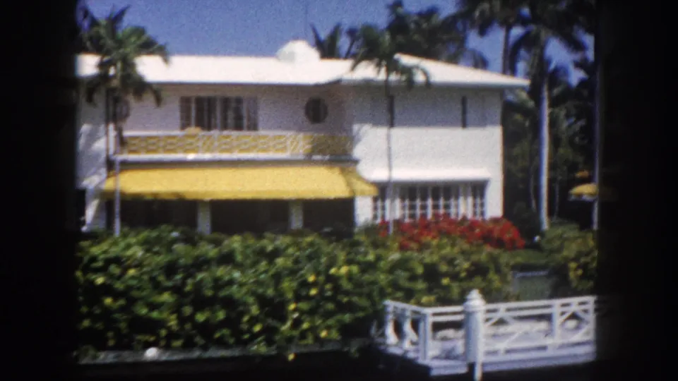 White Florida residence with yellow awning, vibrant red tropical flower beds, and white lattice fence in foreground, Tampa, circa 1960, 8mm.
