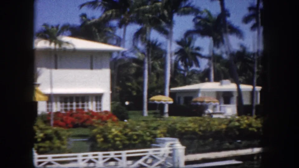 White Florida residence partially screened by palms, with smaller outbuilding and patio umbrella at right suggesting a pool or garden area, Tampa, circa 1960, 8mm.