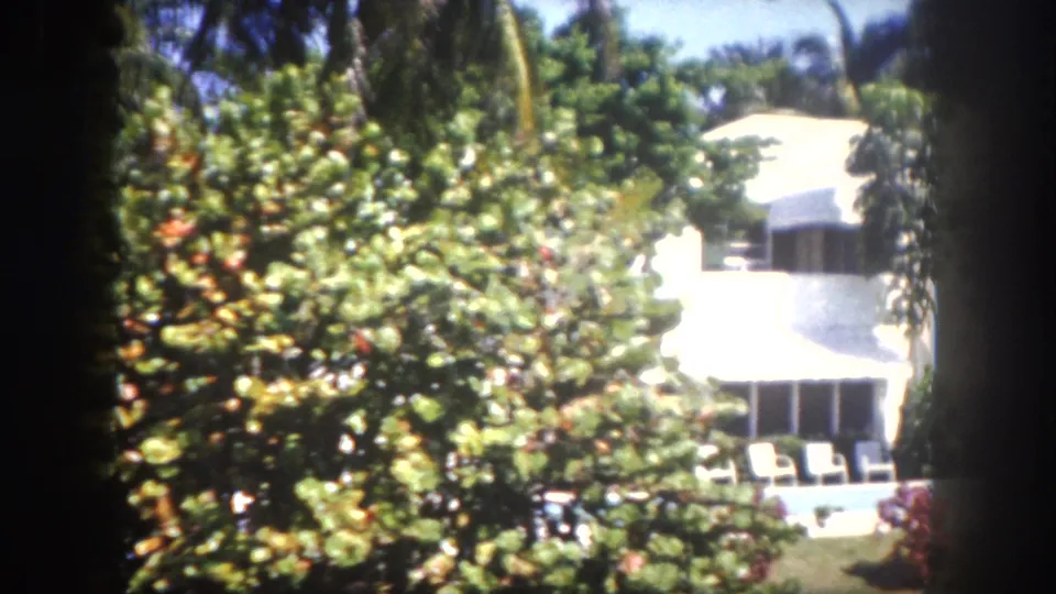 8mm home movie, 1960, Tampa, Florida — white residential house with backyard pool, white patio chairs, and dense red and orange tropical flowering shrubs.