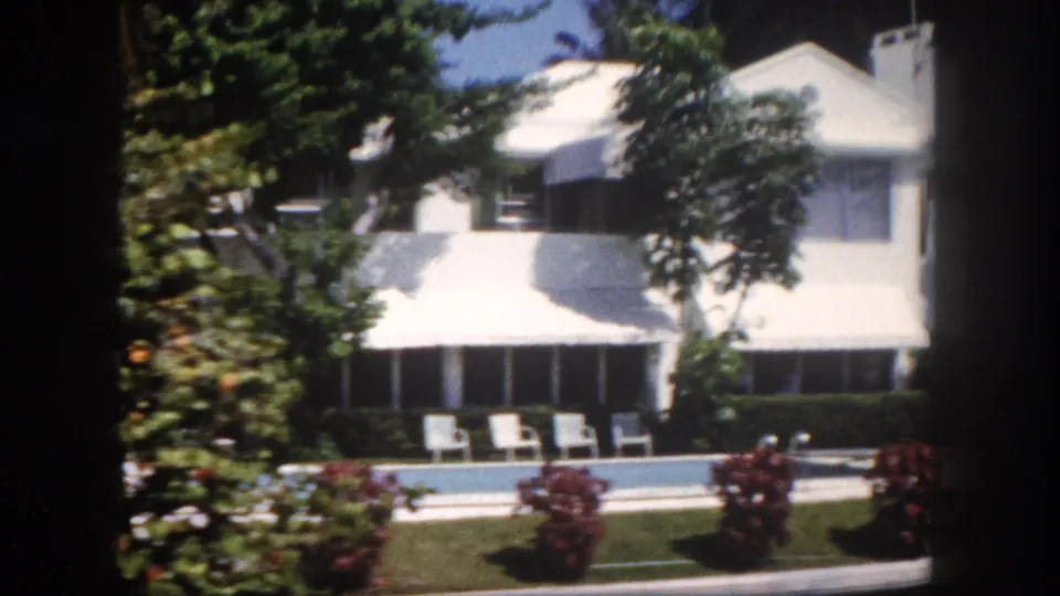 8mm home movie, 1960, Tampa, Florida — large white residential house with screened sunroom, backyard pool, white outdoor chairs, and red tropical flowering plants.