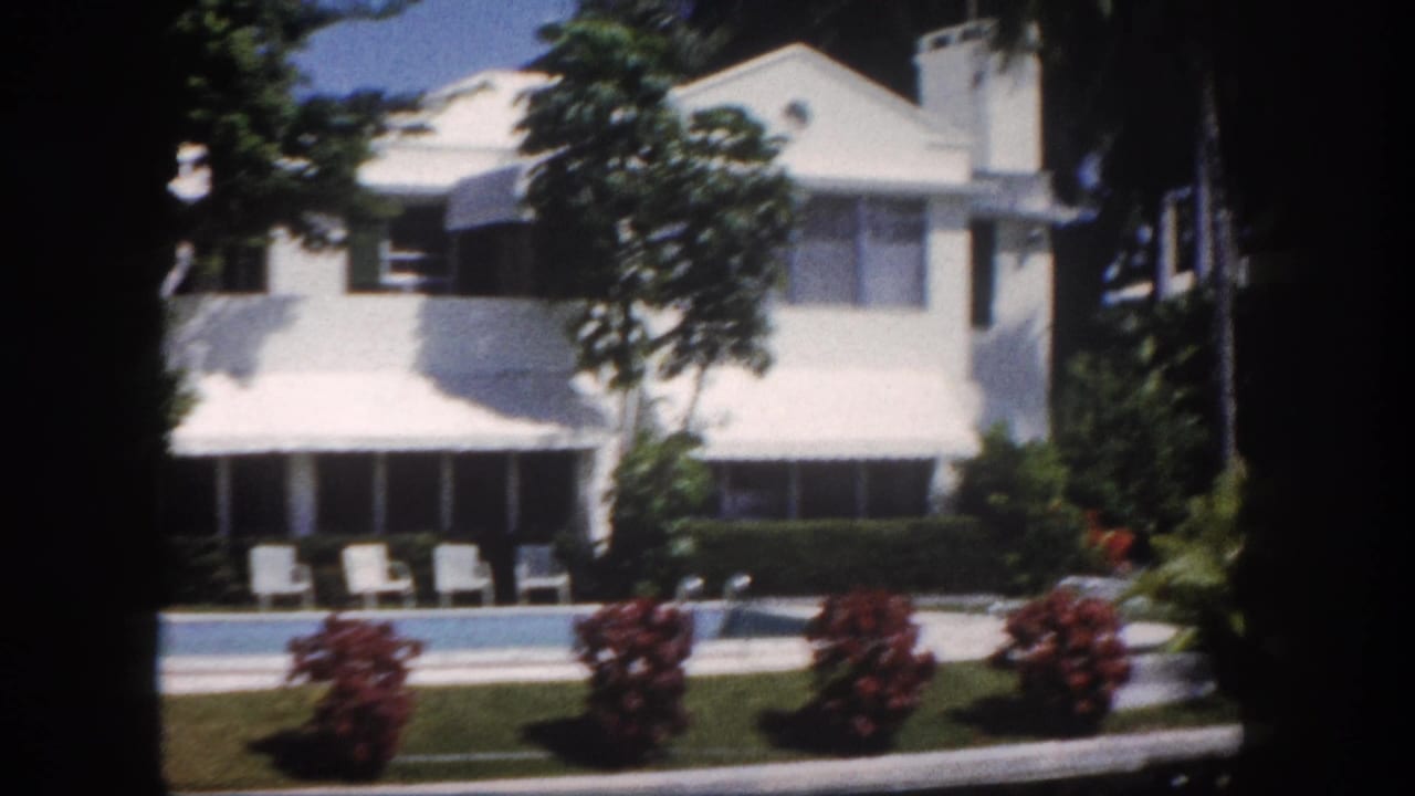 White two-story stucco estate with red tropical flowering shrubs and a swimming pool with lounge chairs in the foreground, palm trees behind.