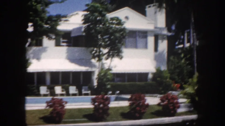1960s 8mm view of a white stucco estate in Tampa, Florida, with red flowering tropical shrubs, a poolside area with lounge chairs, and palm trees.