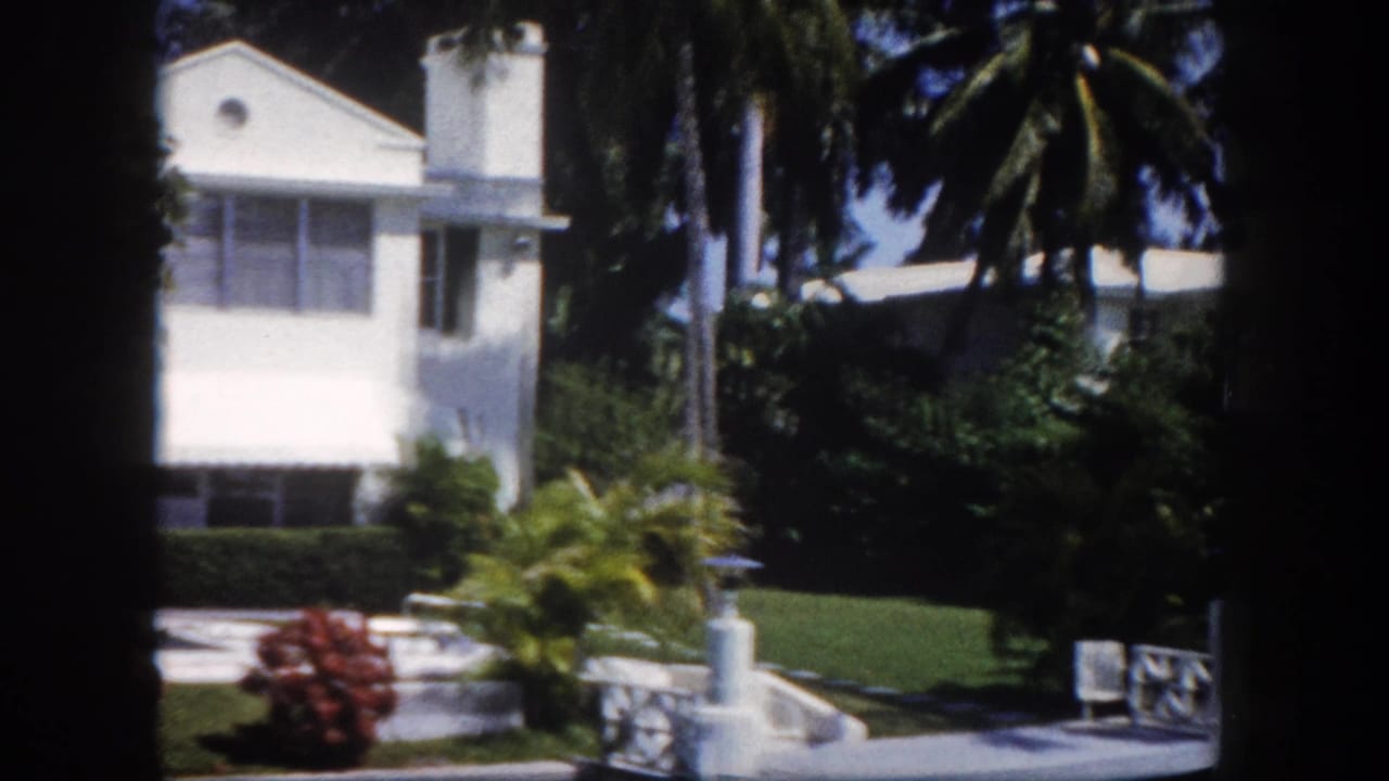 8mm footage from 1960 of a white Florida estate exterior, with red tropical shrubs, tall palm trees, a white garden ornament, and large upper-floor windows.