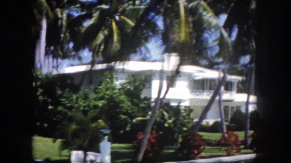 1960s 8mm view of a white estate in Tampa, Florida, framed by tall palm trees, with a white canvas awning and red tropical shrubs visible.