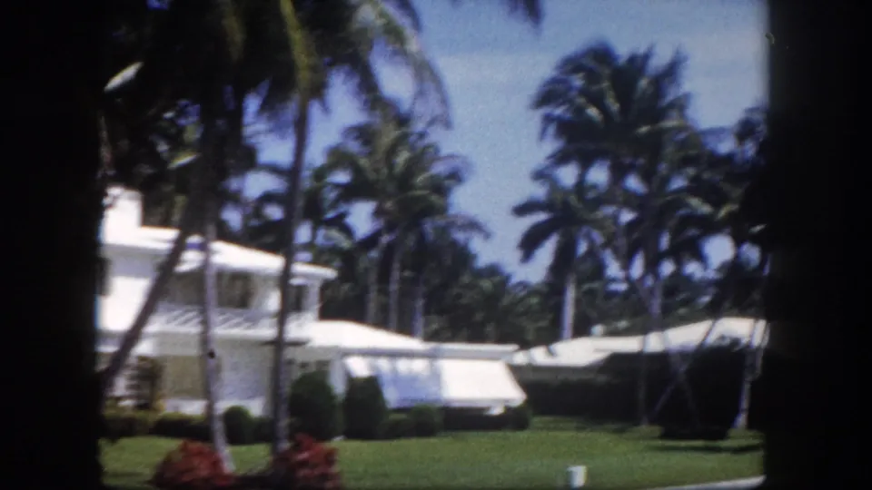 8mm footage from 1960 showing a white Florida estate and canvas awning framed by mature palm trees, with red tropical shrubs on the foreground lawn.