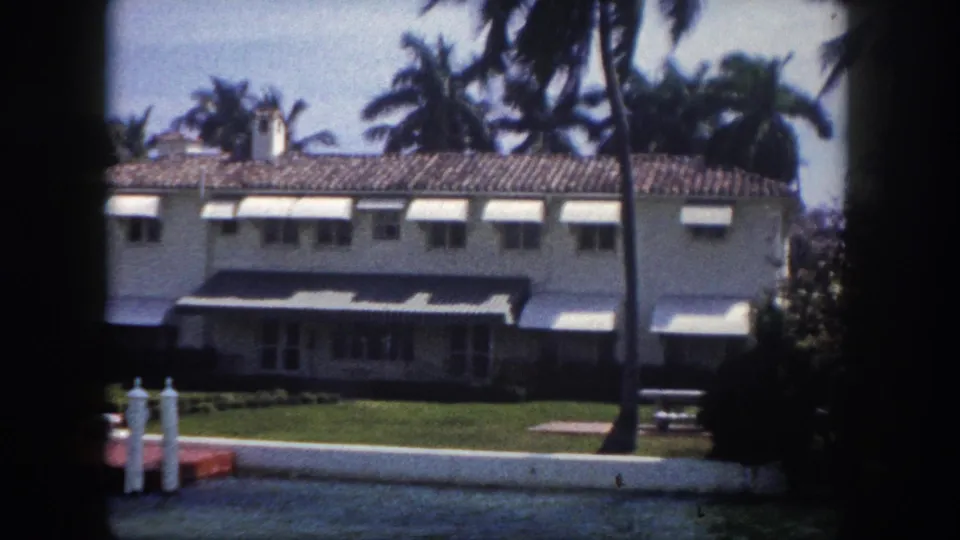 Mid-century white house with striped canvas awnings and palm trees along a Florida canal, circa 1960, with dock and mooring bollards in foreground, 8mm home movie.