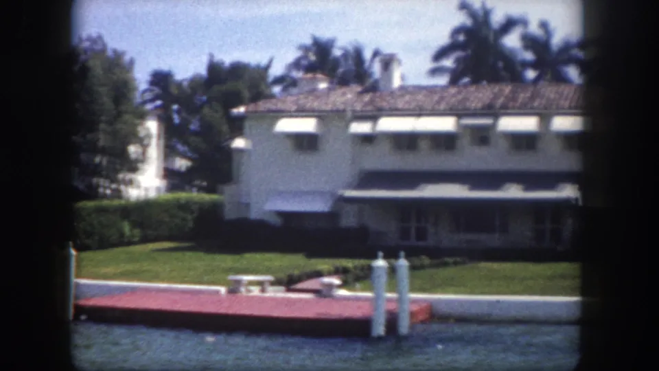 Two white mid-century waterfront homes with striped canvas awnings and mooring dock along a Florida canal, circa 1960, 8mm home movie from a passing boat.