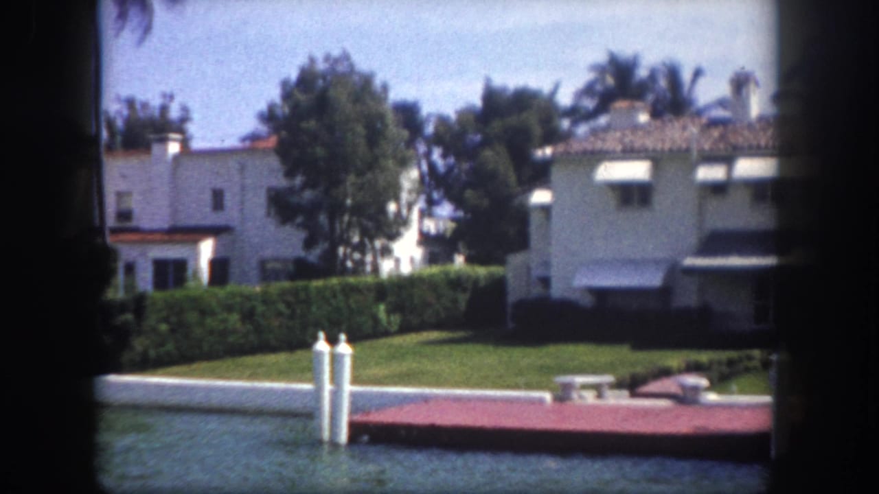8mm footage circa 1960, Tampa, Florida — white stucco waterfront homes with terracotta roofs, green lawn, tall palm trees, and white dock pilings in foreground.
