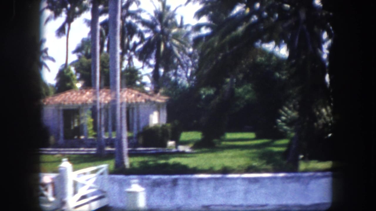 8mm footage circa 1960, Tampa, Florida — white stucco Mediterranean home with terracotta roof, manicured lawn, palm trees, and low white balustrade fence at water's edge.