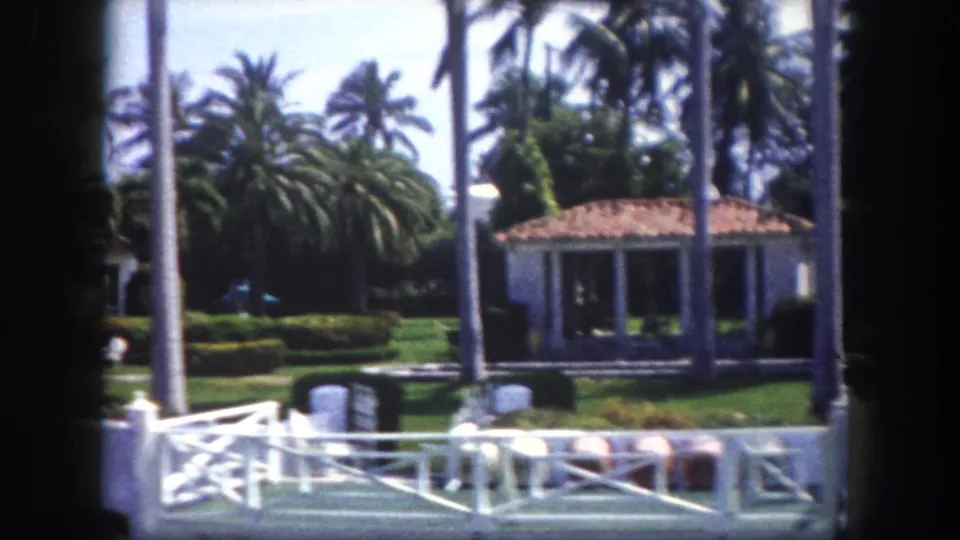 8mm footage circa 1960, Tampa, Florida — white columned open pavilion beside a Mediterranean home, dense palm trees, manicured hedges, and white balustrade in foreground.
