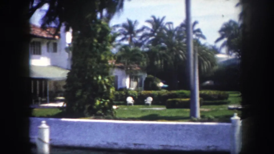 8mm footage circa 1960, Tampa, Florida — two-story white stucco home with terracotta tile roof, dense palm canopy, tropical garden, and white balustrade fence along waterfront.