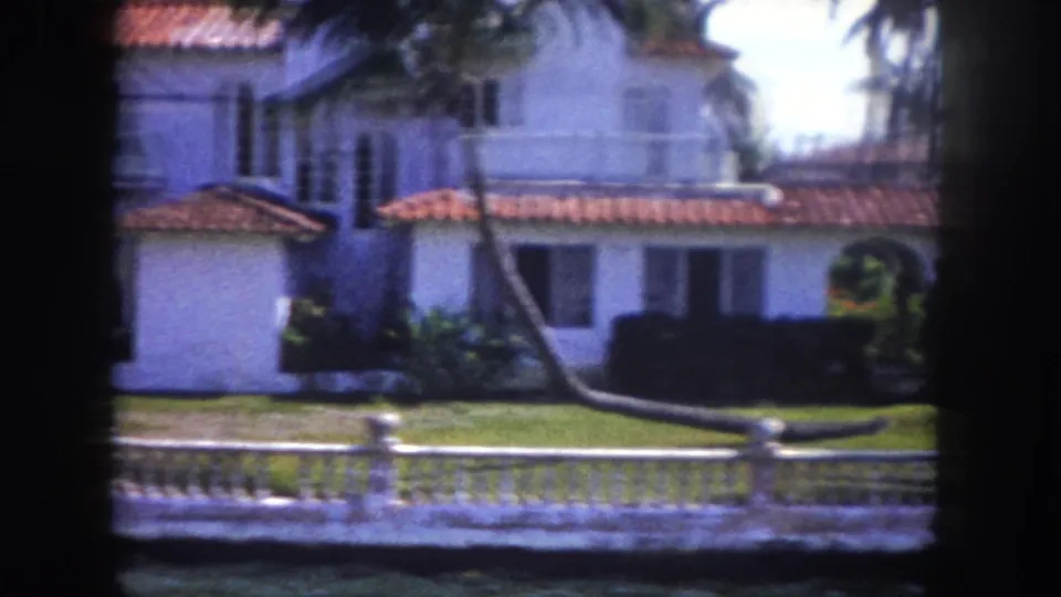 Single-story white stucco waterfront home with terracotta tile roof and ornamental fence filmed from a Tampa canal, circa 1960.