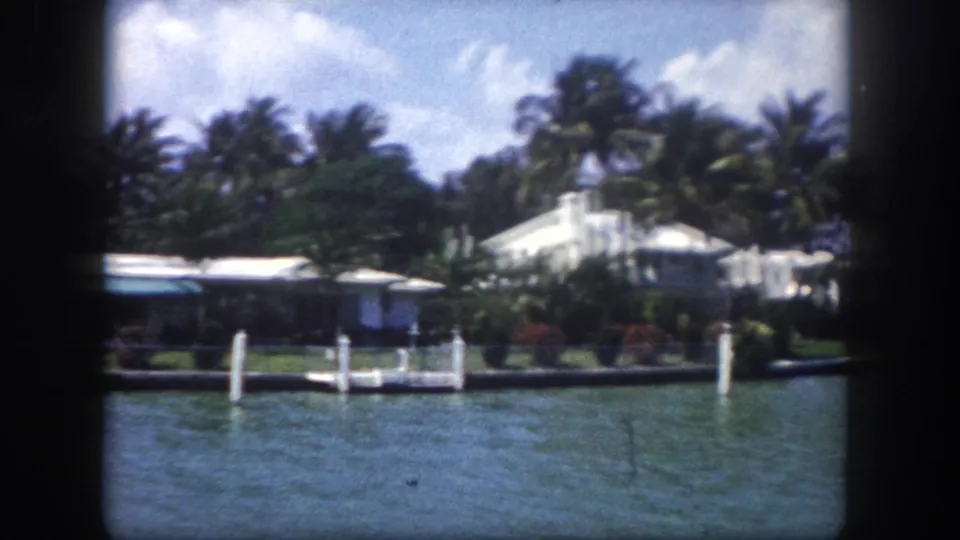 1960s Tampa, Florida: clearest frame shows a white waterfront estate with dock pilings in the foreground and dense palm trees behind.